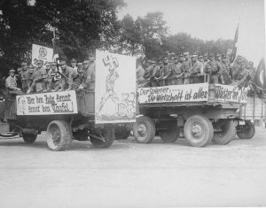 Members of the SA drive through the streets of Recklinghausen, Germany with anti-Jewish banners during a propaganda parade. One banner reads, “He who knows the Jew knows the Devil.” The banner in front depicts a German man using a swastika to crush Jews who are shown with snakes coming out of their heads. August 18, 1935. Photo credit: USHMM #80821