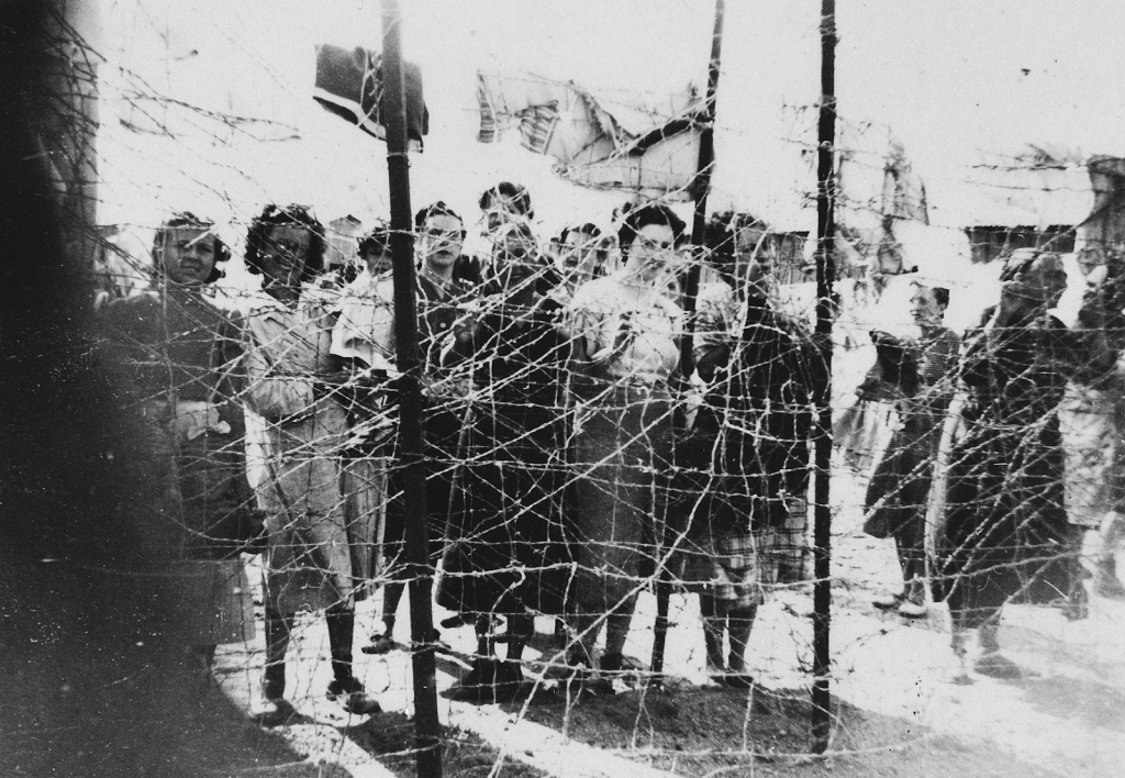 Female prisoners gather at the barbed wire fence surrounding the Gurs concentration camp in Southern France, circa 1939-1940. Photo credit: USHMM #65543