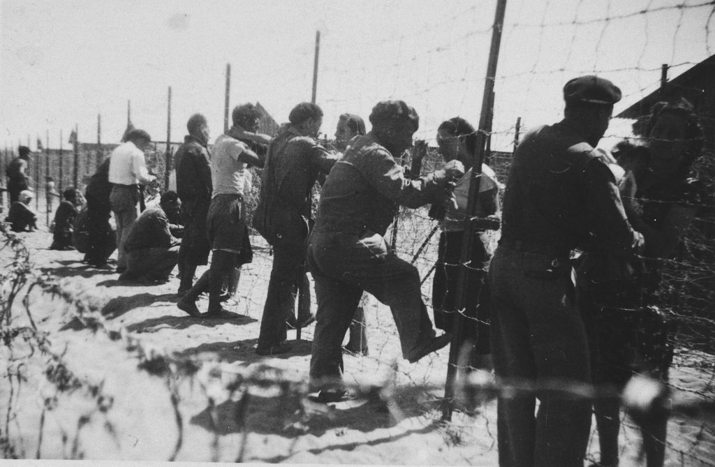 Prisoners gather by the barbed wire fence surrounding the Gurs internment camp.