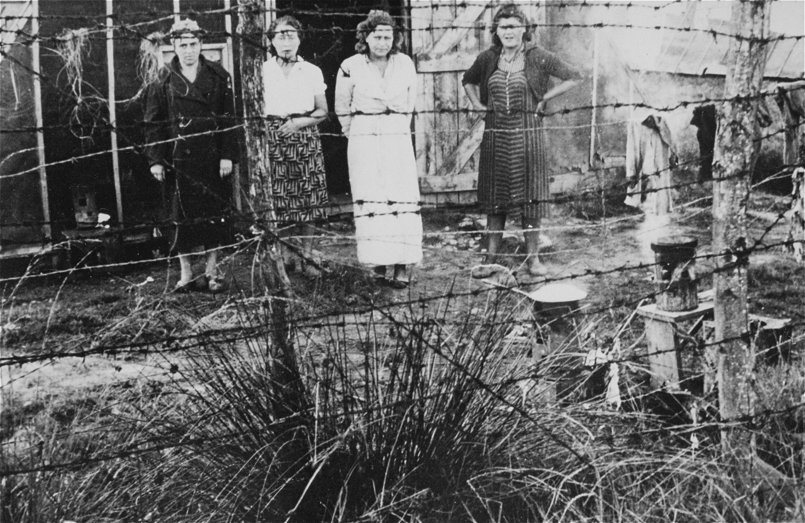 Four female prisoners stand outside a barracks behind a barbed-wire fence in 
